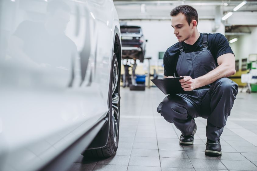 Mechanic in overalls kneeling next to white car in garage, carrying out maintenance checks