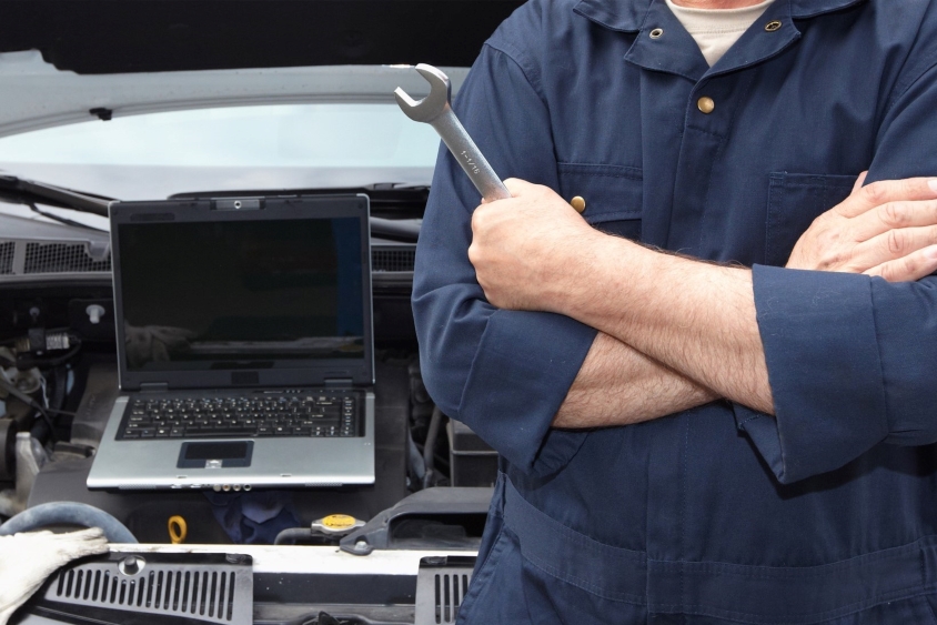 Mechanic with arms crossed holding spanner next to a car engine and a laptop