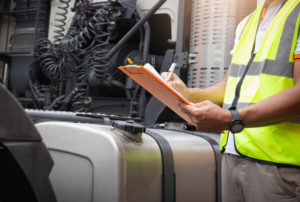 A truck driver holding clipboard checking safety a large fuel tank of semi truck.