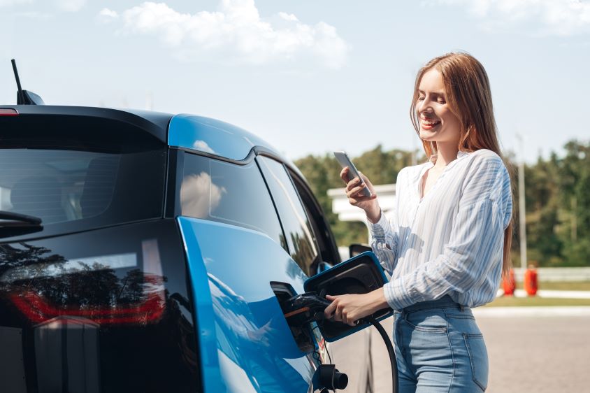 Woman standing next to blue car whilst it charges, looking at her phone